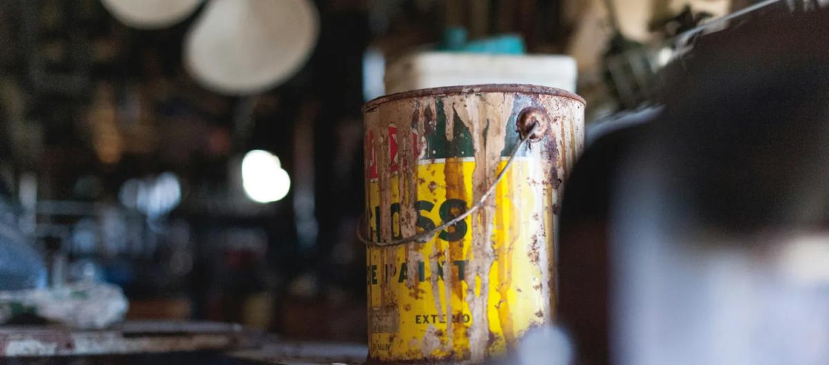 Rusty and paint-splattered can of gloss exterior paint sitting in a dimly lit, cluttered garage.